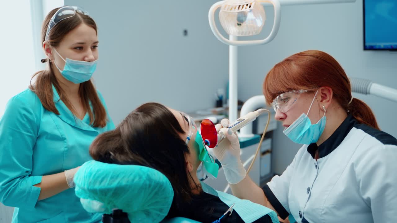 Female stomatologist and a nurse treating woman's teeth. Dentist working in patient oral cavity. Woman sitting in the dental chair at teeth treatment in dental office.
