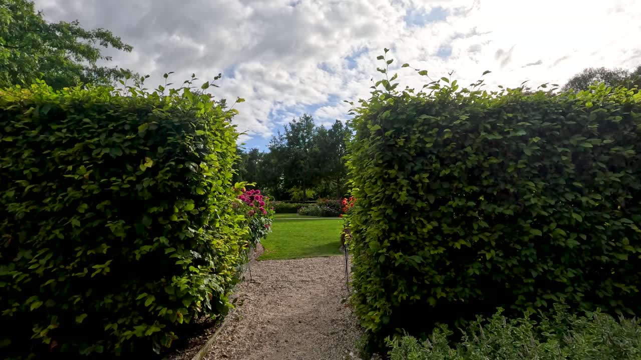 A steady camera moves along a gravel path beside manicured hedges and blooming flowerbeds in a sunlit botanical garden, with soft natural lighting and lush greenery