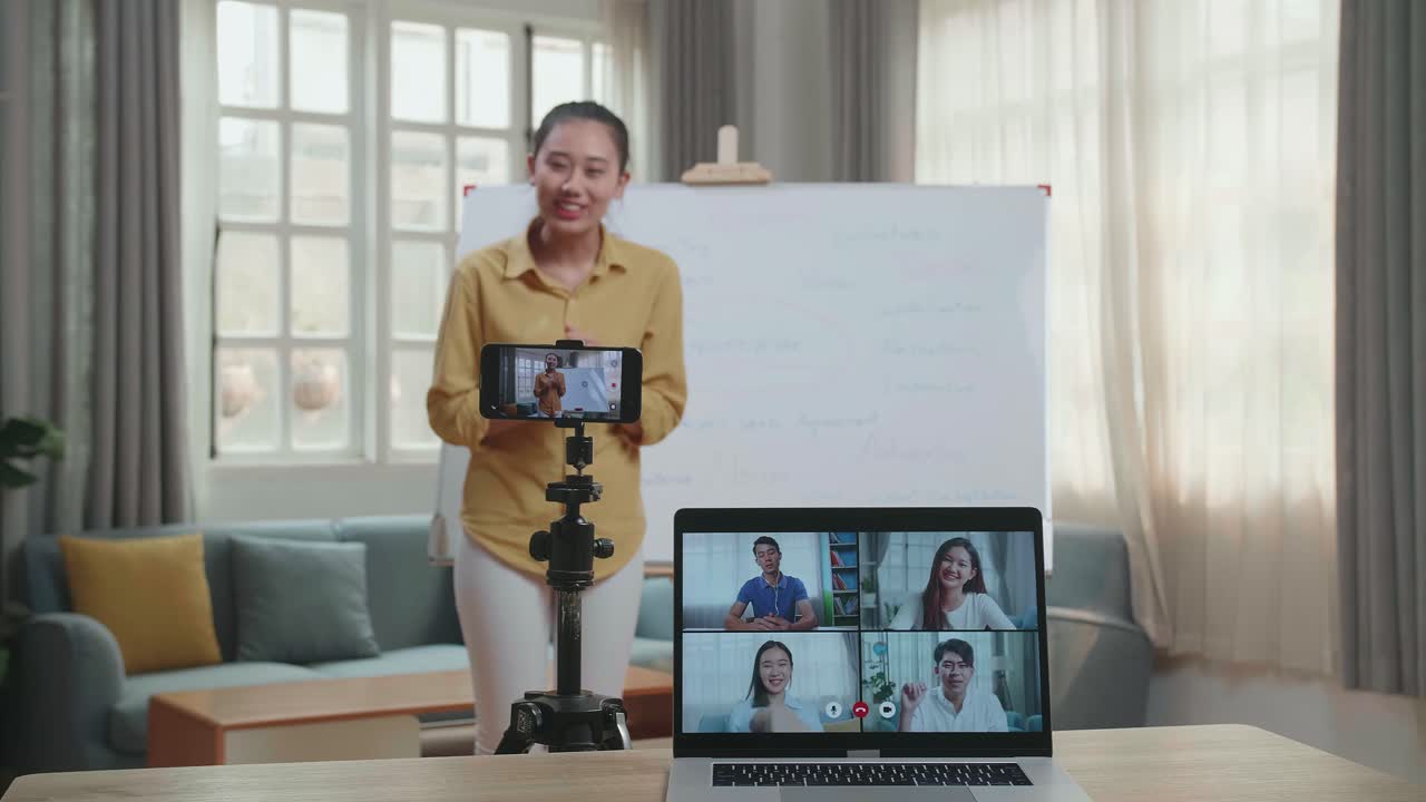Asian Female Teacher Shoots Video By Smartphone Camera And Having Video Call On Laptop While Teaching English At Home