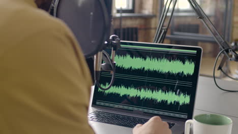 vista traseira de um homem sentado em uma mesa ajustando o equalizador no laptop durante a gravação de um podcast