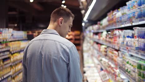 Und-Attraktiver-Großer-Kerl-Im-Supermarkt.-Seltener-Blick-Auf-Kaukasischen-Jungen-Mann-Im-Blauen-Hemd-Auf-Der-Suche-Nach-Einer-Sauren-Sahne-Und-Nehmen-Sie-Es-Aus-Dem