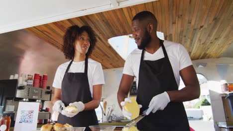 Pareja-Afroamericana-Usando-Delantales-Sonriendo-Mientras-Preparan-Perros-Calientes-Juntos-En-El-Camión-De-Comida