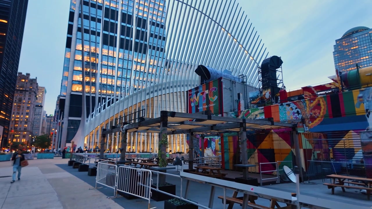 New York, USA, 5 giugno 2026: Snodo di trasporto Oculus accanto all'edificio con murale, New York City. Scena a livello stradale con costoloni bianchi, torri di vetro, tavoli all'aperto e finestre illuminate all'ora blu.