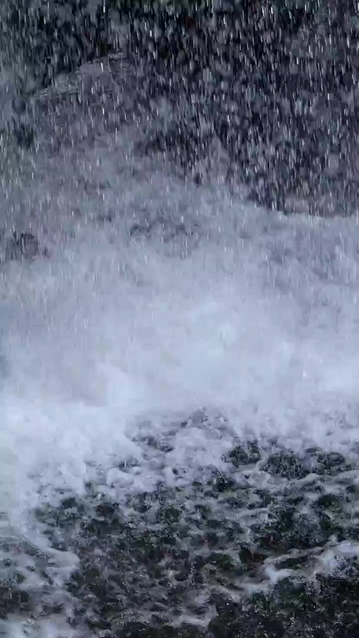 Air terjun bergejolak menghantam bebatuan gelap, menciptakan percikan dan kabut yang intens. Pencahayaan alami, perspektif jarak dekat, dan gerakan dinamis menonjolkan kekuatan mentah dan tekstur.