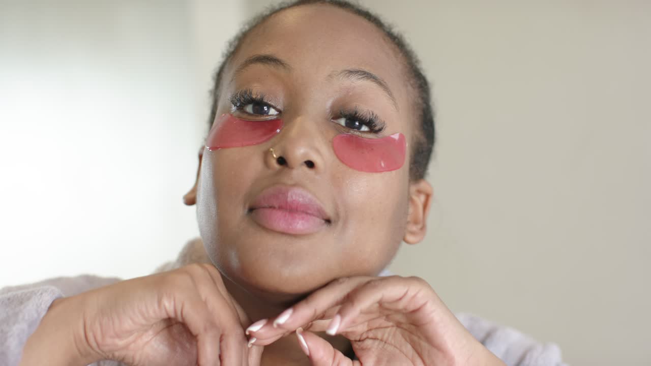 Starting skincare African American woman applying, massaging eye gel patches at bathroom vanity