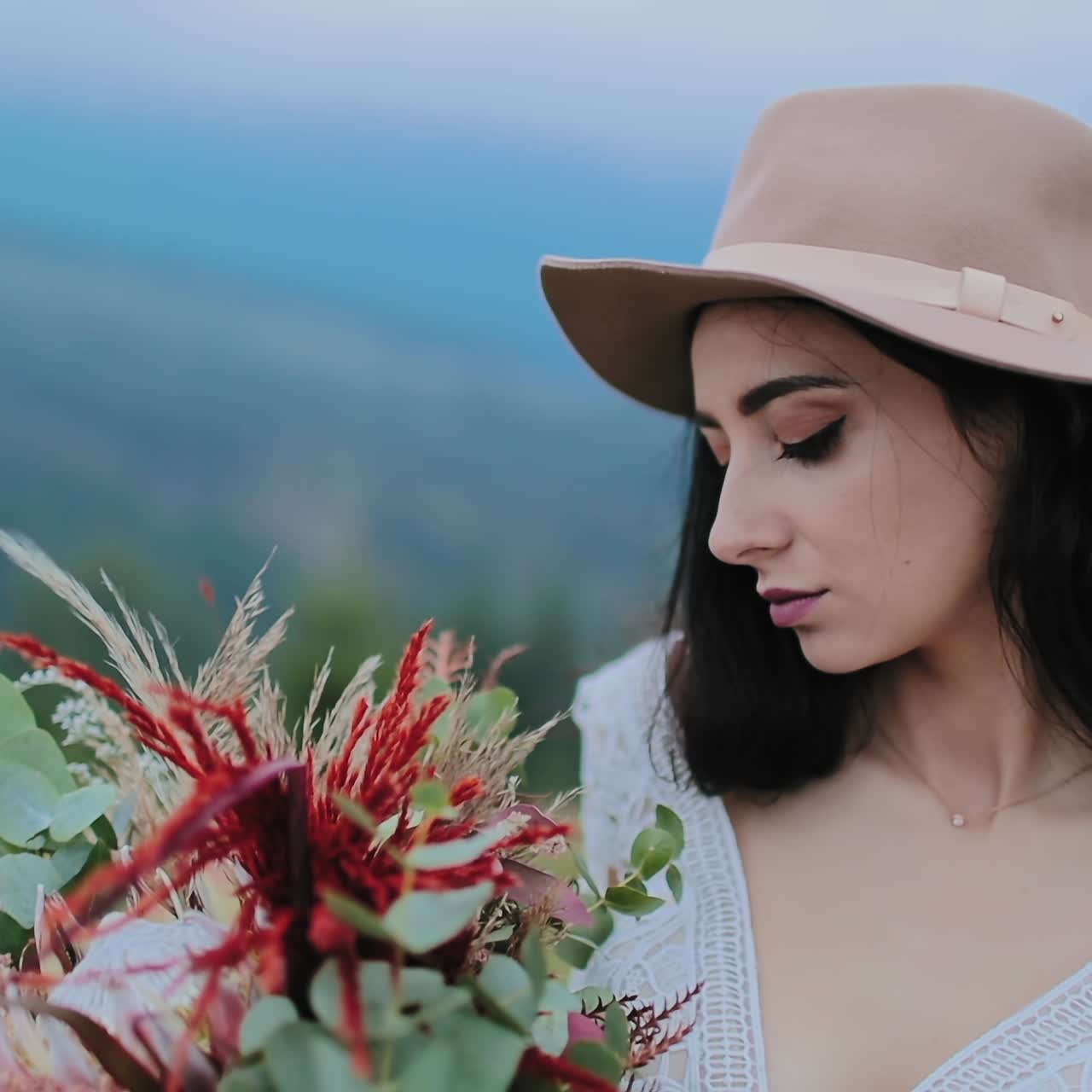 Beautiful girl with wild flowers bouquet. Young woman in hat looking at flowers in her hands on the background of mountains. Close-up.