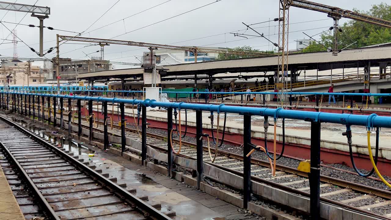 Platform view at Vadodara railway station, railway tracks with central waterline as train approaches under cloudy sky in Gujarat, India.