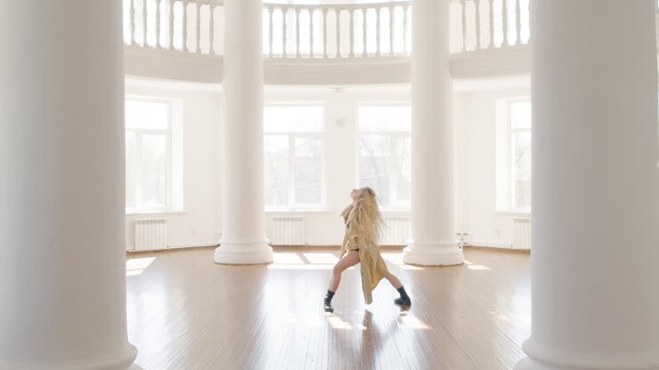 Focused Blonde Woman In Trench Coat And Boots Training A Contemporary Dance In The Middle Of The Studio