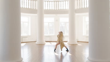 mujer rubia enfocada en gabardina y botas entrenando una danza contemporánea en medio del estudio