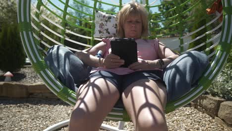 woman reading ebook novel relaxing in comfortable garden swing hammock on hot sunny day