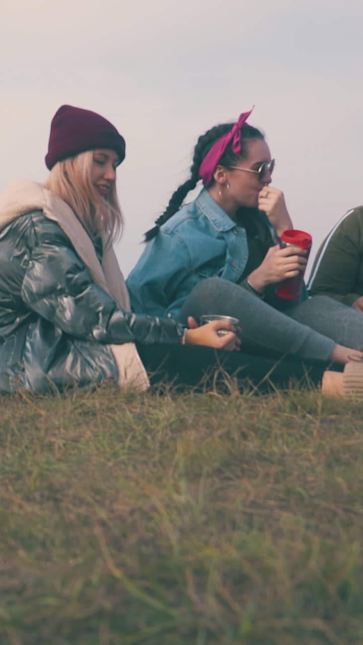 smiling young women with drinks enjoy time sitting on grass at tent against clear sky in autumn evening low angle shot