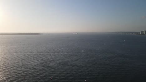 Cargo ship navigating calm waters near urban coastline during sunrise, creating a serene maritime scene