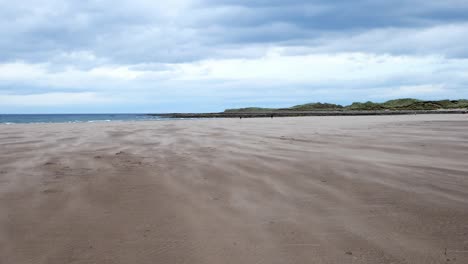 Handheld-slow-motion-shot-of-wind-patterns-of-sand-blowing-across-the-flat-beach-on-the-Holy-Island,-Lindisfarne,-England