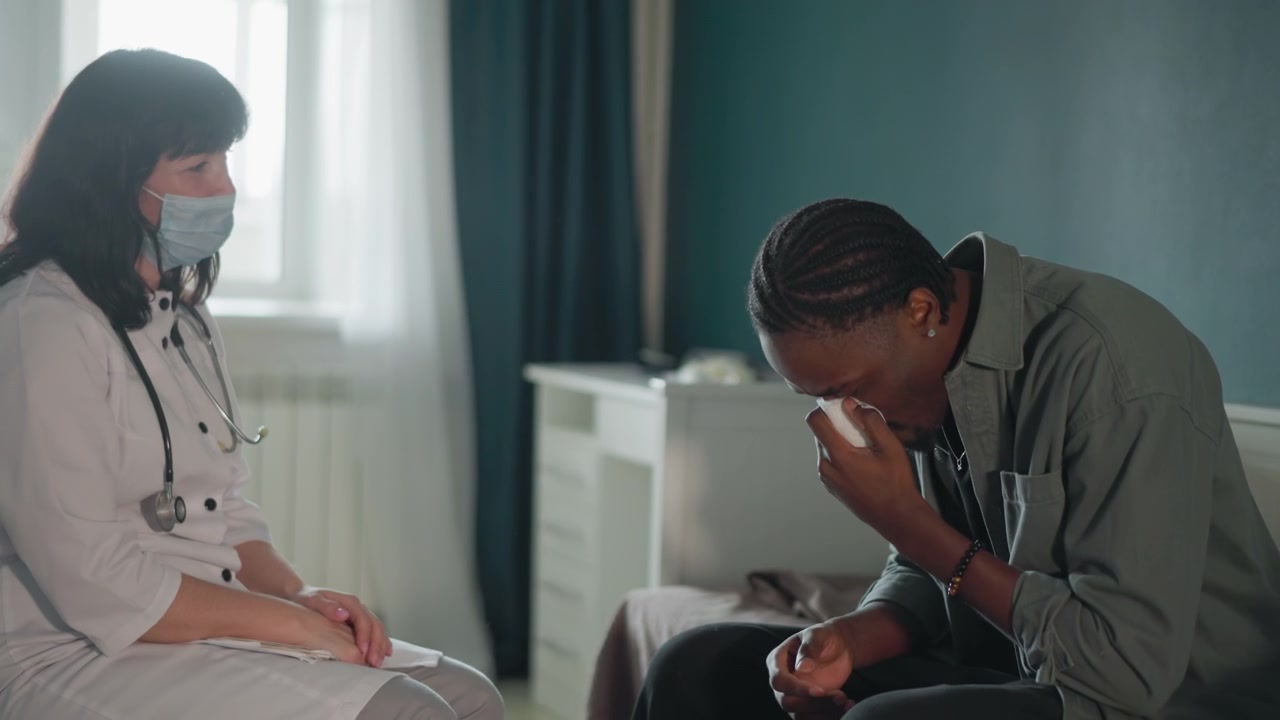 Svart mann sitter fortvilet på sengen, hvit lege med maske og stetoskop tilbyr rolig støtte og veiledning til tårevåte hender, papirlommetørkle i nærheten, rolig soveromsmiljø med mykt vinduslys