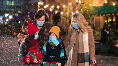 nahaufnahme einer kaukasischen glücklichen familie mit gesichtsmasken, die die straße hinuntergeht und geschenke hält, während es zu weihnachten schneit