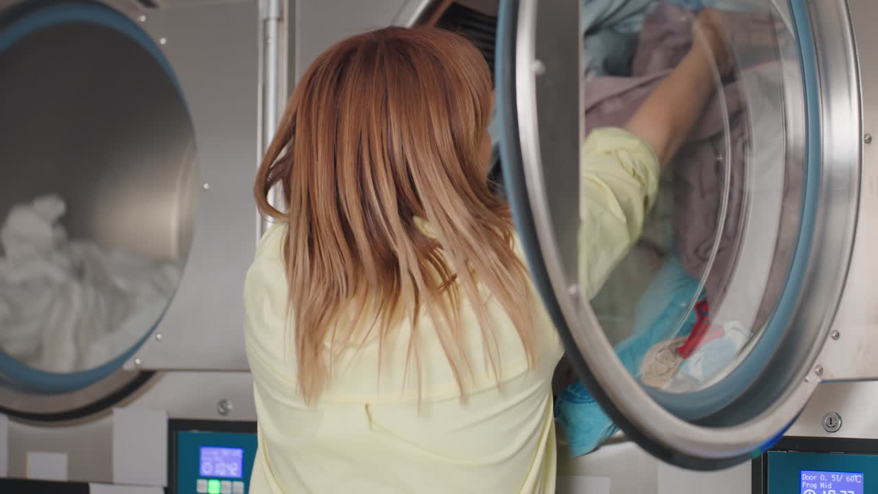 Back view of laundry owner carrying pile of clothes, transferring load from washer into upper dryer compartment, closing machine door while fabrics roll inside steel equipment with digital display