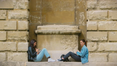 two female university or college students sitting against wall in city working on digital tablet