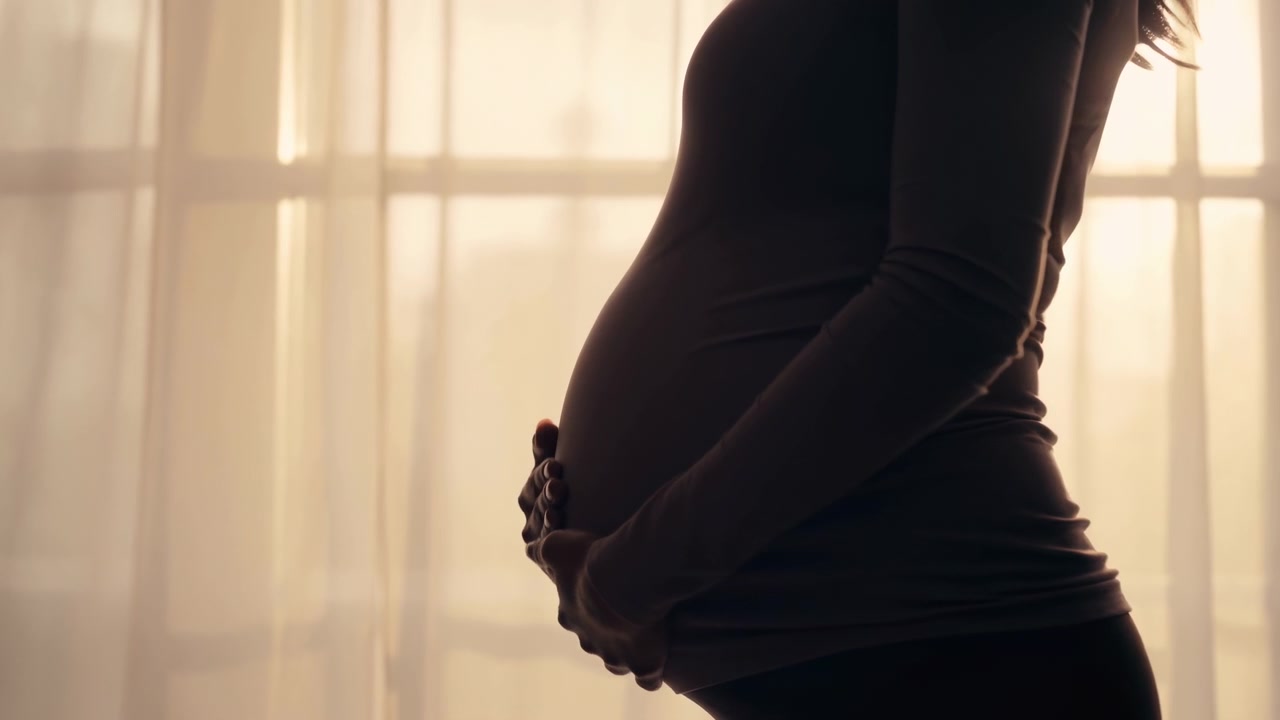 Sidebilde av en gravid kvinne som forsiktig holder magen sin nær et solfylt vindu med tynne gardiner, noe som skaper en rolig, intim hjemmeatmosfære med varmt bakgrunnslys.