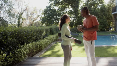 happy diverse couple holding yoga mats talking in sunny garden, copy space, slow motion