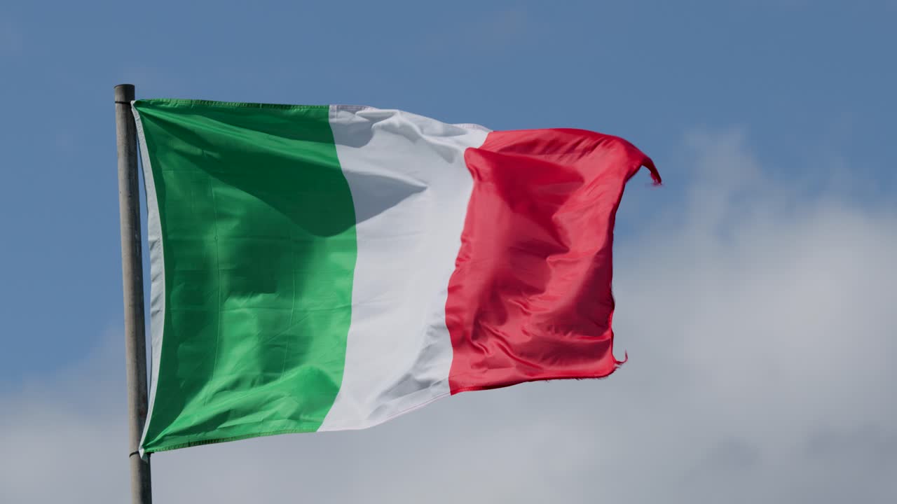 La bandiera tricolore italiana sventola dinamicamente su un pennone, luce diurna intensa, cielo sereno, telecamera stabile