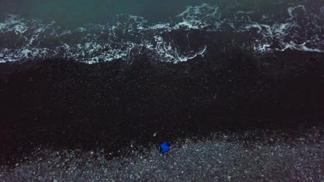 Aerial-rising-top-down-view-of-boy-throwing-volcanic-pebble-stones-into-sea