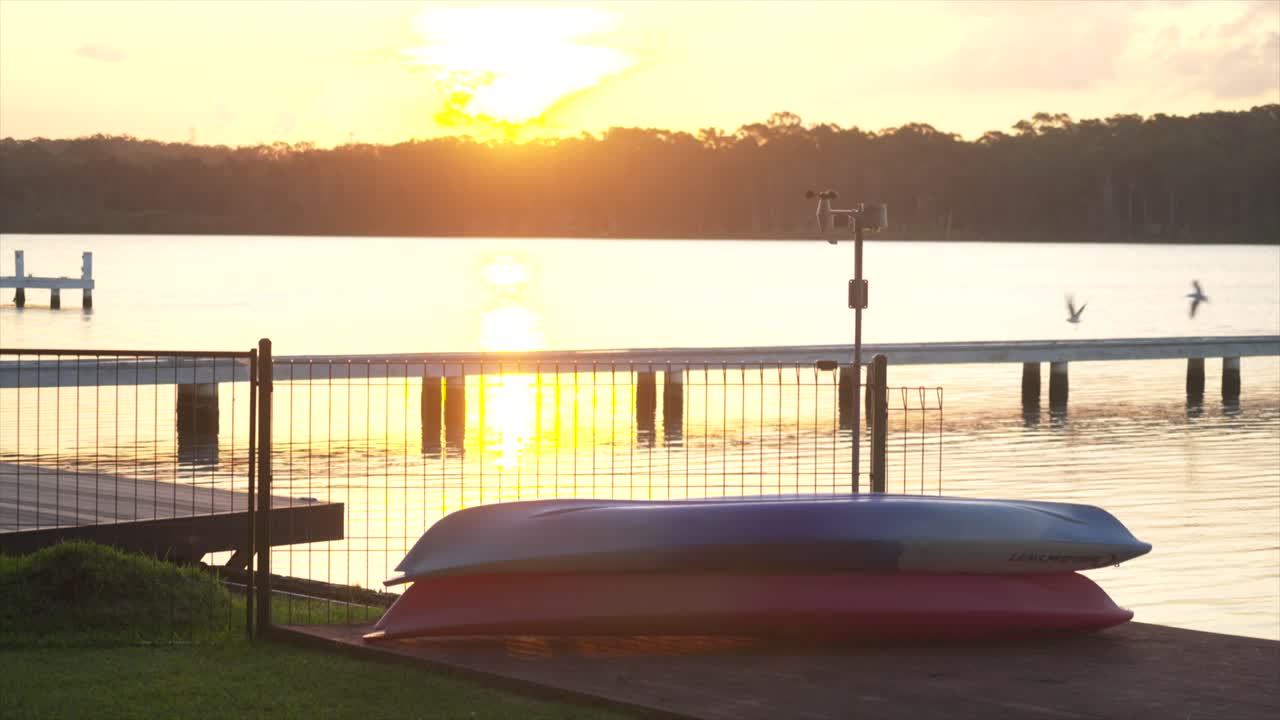 Kayak impilati sulla riva con il tramonto sul Lago Macquarie e un pontile sullo sfondo, Nuovo Galles del Sud, Australia