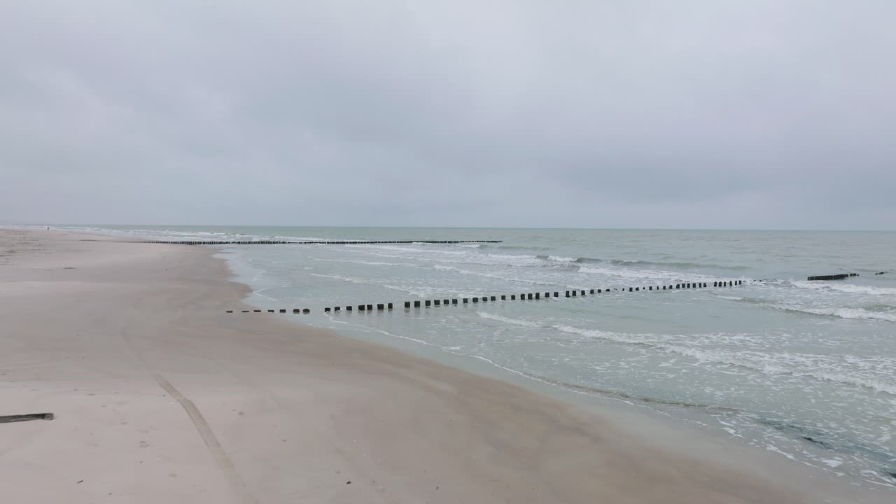 발트해 연안에 있는 오래된 목조 부두의 공중 전망, 흐린 겨울날, 하얀 모래 해변, 나무 기둥, 해안을 강타하는 파도, 앞으로 낮게 움직이는 넓은 드론 샷