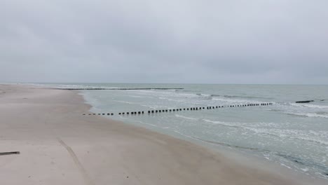 Aerial-establishing-view-of-an-old-wooden-pier-at-the-Baltic-sea-coastline,-overcast-winter-day,-white-sand-beach,-wooden-poles,-waves-hitting-the-shore,-wide-drone-shot-moving-forward-low