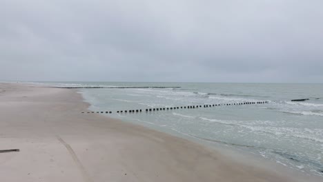 aerial establishing view of an old wooden pier at the baltic sea coastline, overcast winter day, white sand beach, wooden poles, waves hitting the shore, wide drone shot moving forward low