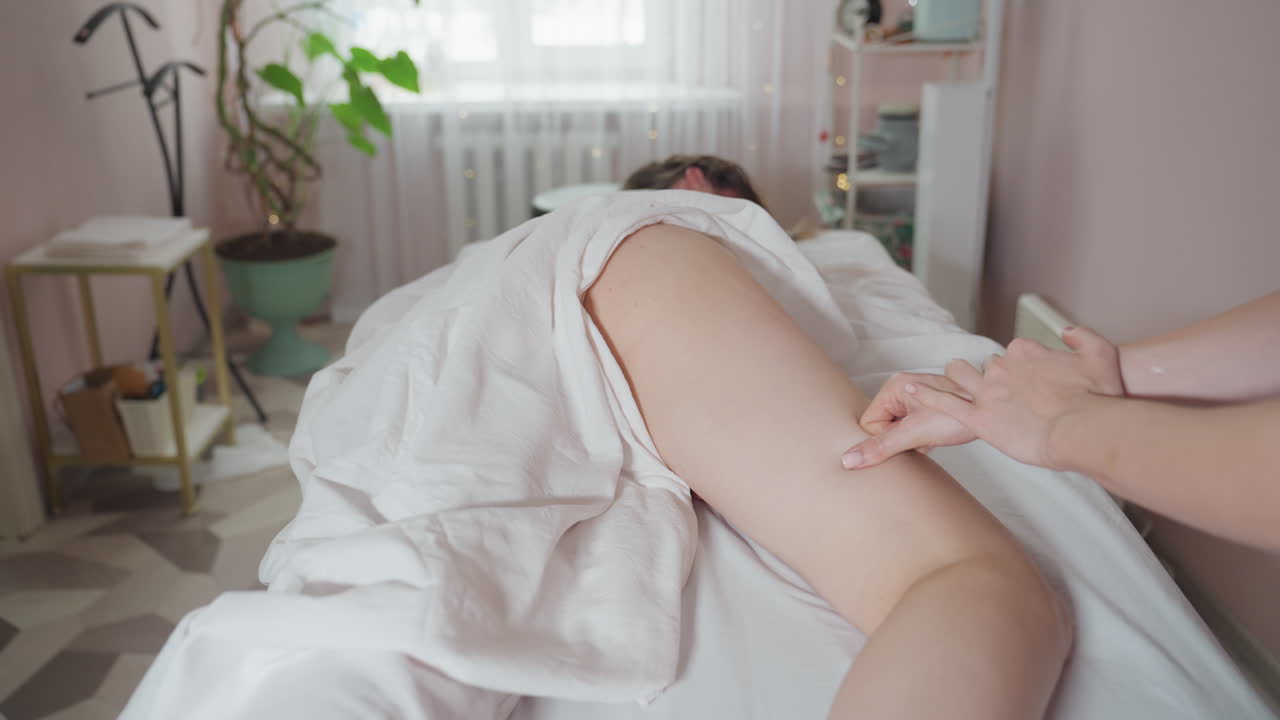 Massage therapist in black attire deeply focused on applying pressure to client s thigh using both hands while client lies under white sheet in softly lit spa room with blurred plant background