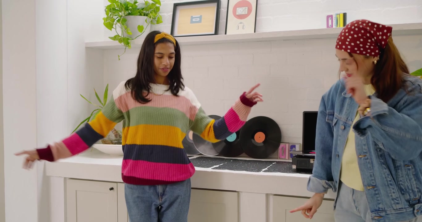 Two women in striped sweater handling vinyl in kitchen, starting turntable music, dancing