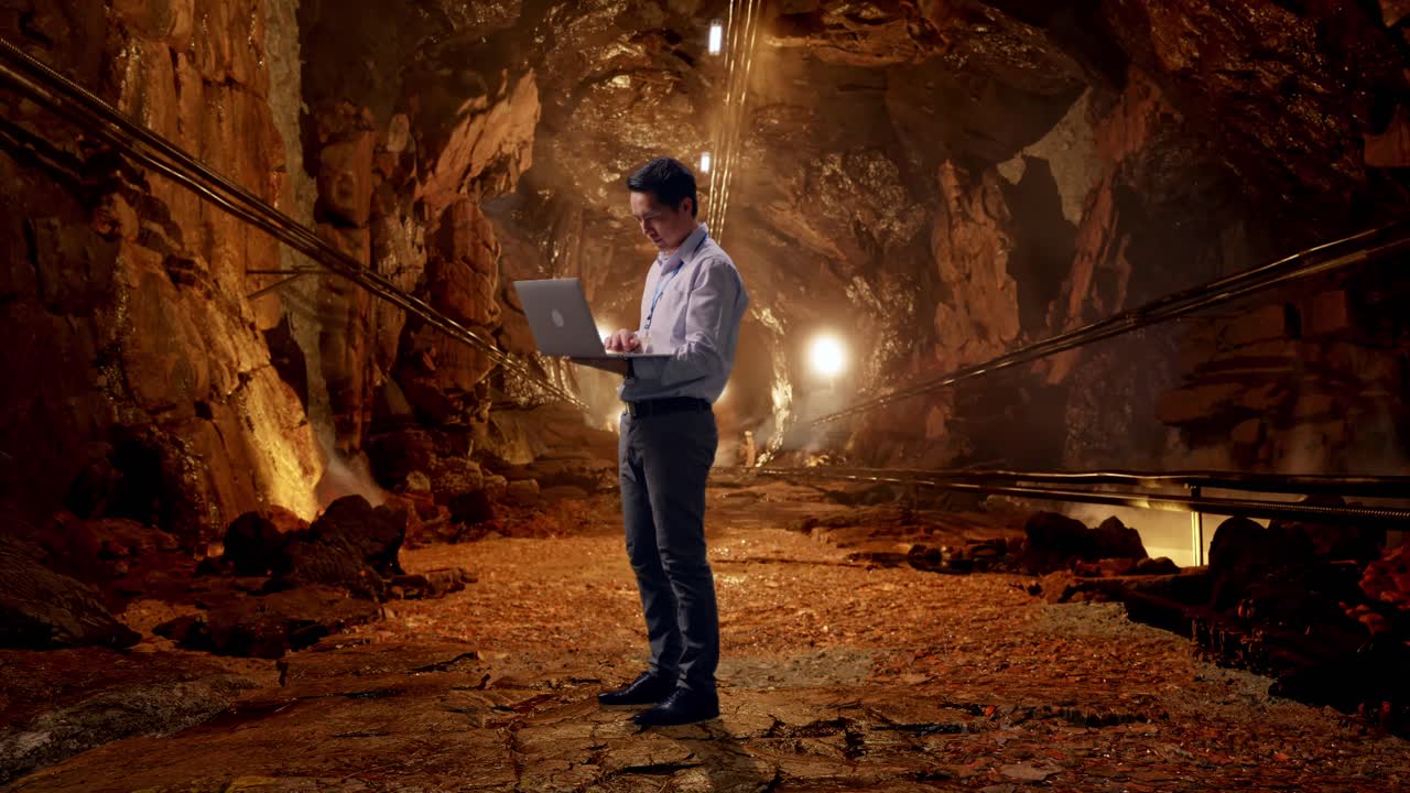 Full Body Side View Of An Asian Male Professional Worker Standing With His Laptop In Underground Mine Tunnel, Typing On His Laptop'S Keybaord With Meditation