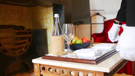 Butler-puts-tray-with-water-bottle-and-a-bowl-of-fruit-in-a-table-next-to-a-fireplace-in-an-antique-building