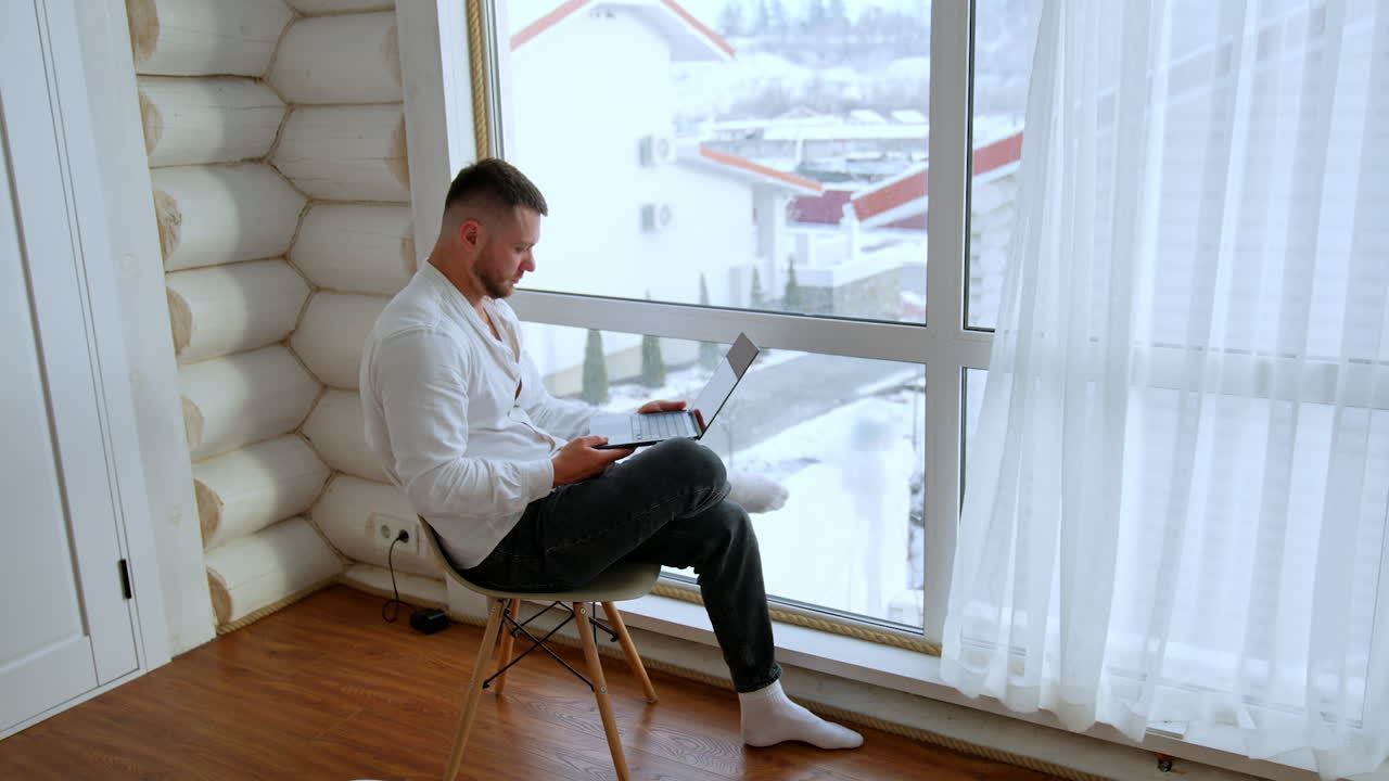 Man sitting on the chair at the window uses his laptop. Freelancer working remotely from home.