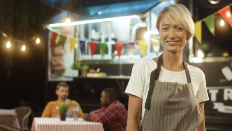 Happy-Beautiful-Joyful-Young-Woman-In-Apron-With-Blonde-Short-Hair-Smiling-At-Festive-Food-Track-In-Evening