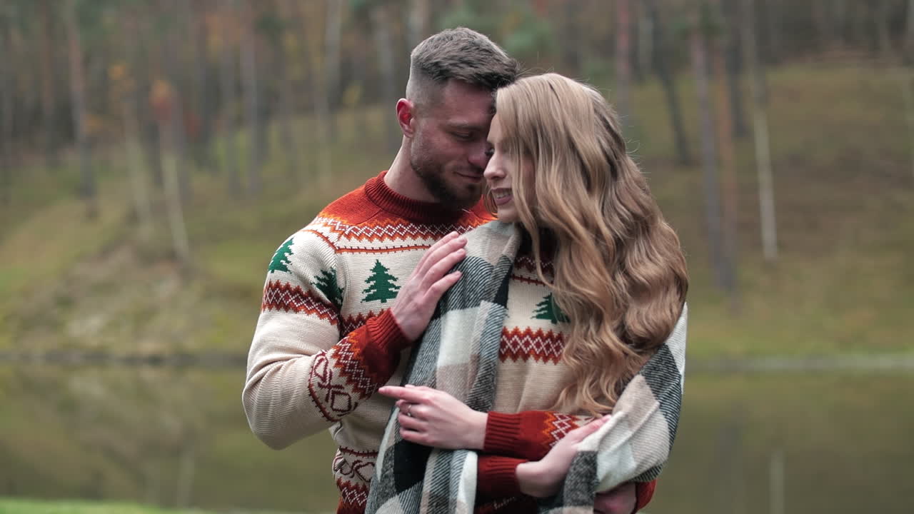 Man caressing his girlfriend covered with warm blanket. Happy loving couple standing near the river. Nature at the background.