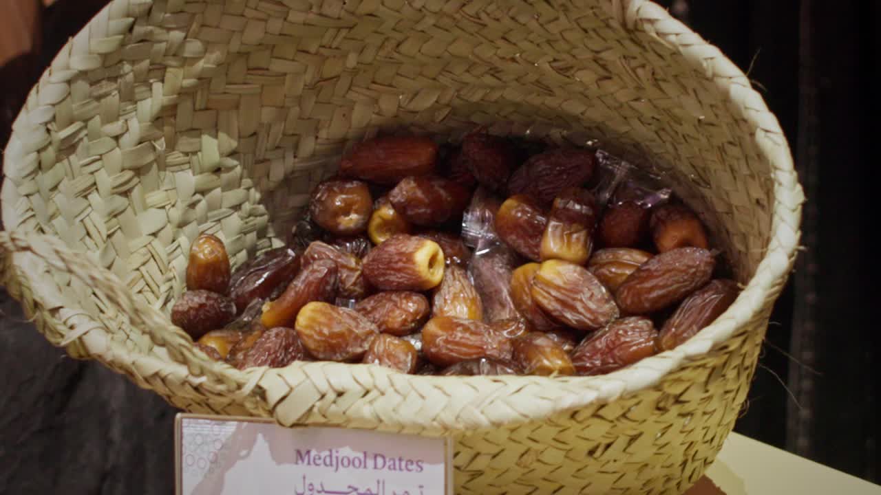 Close-up of fresh Medjool dates piled inside a traditional woven basket at a market stall. The rich brown dates are glossy and ripe, presented in a rustic