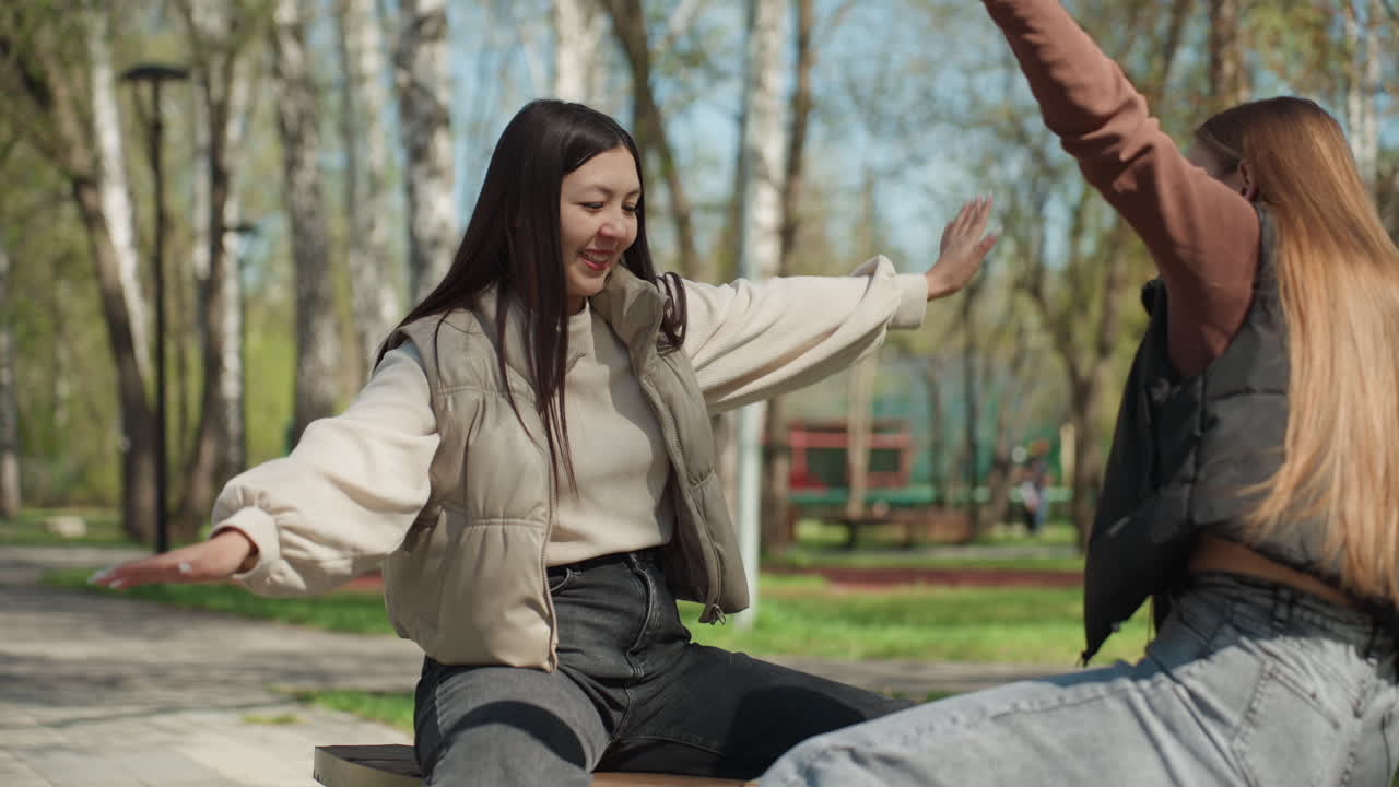 Две женщины улыбаются, сидя на скамейке в парке. Женщины азиатской и европейской внешности наслаждаются спонтанной радостью на открытом воздухе. Подруги разного происхождения вместе переживают счастливые моменты на свежем воздухе.