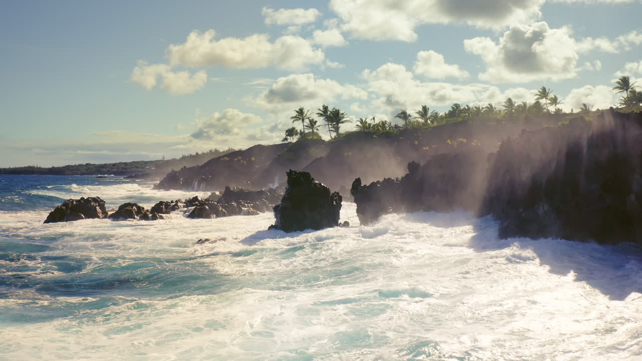 Thunderous ocean waves crash against jagged volcanic cliffs beneath a palm-dotted shoreline, their white spray rising into the sunlight as the Pacific displays its untamed power