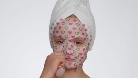 Lovely-young-smiling-child-girl-applying-cosmetic-moisturizing-face-mask,-use-massage-roller