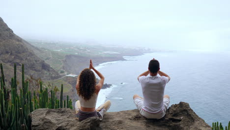 Atop-a-mountain,-a-man-and-woman-sit-on-stones,-meditating,-their-hands-raised,-as-they-look-out-over-the-ocean-and-perform-calming-breath-exercises