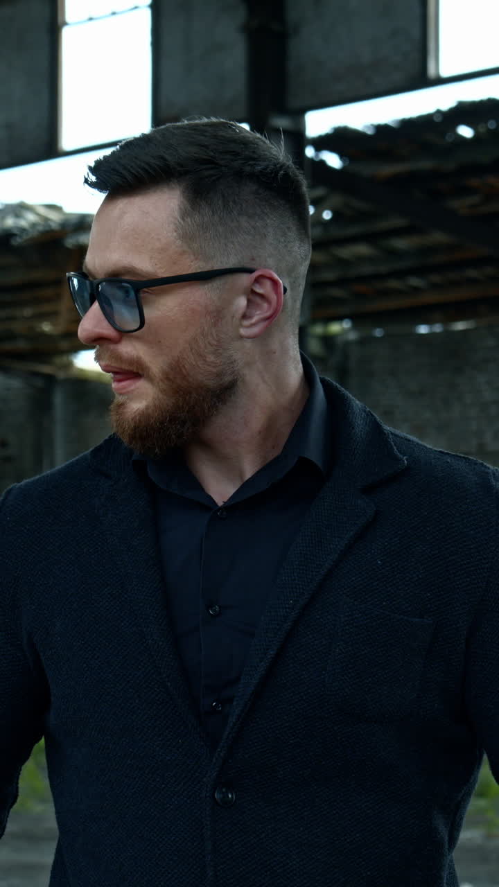 Elegant man with a baseball bat outdoors. Portrait of a serious man in stylish glasses on the background of abandoned building. Vertical video