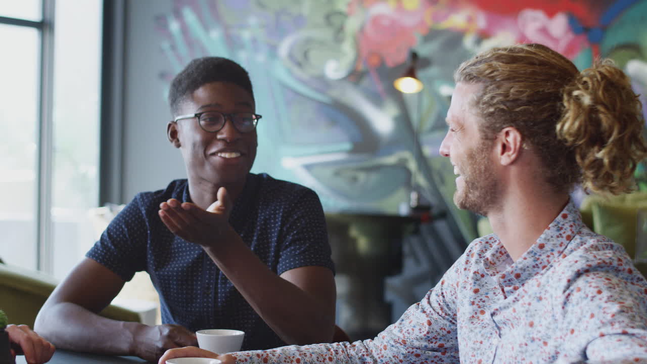 Two happy young male creatives talking with colleagues in their workplace canteen, close up