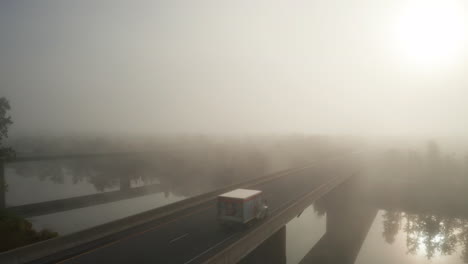 Peligrosas-Condiciones-De-Conducción-Nublada-Con-Automóviles-Y-Camiones-Que-Conducen-Por-Un-Puente-De-Carretera-Que-Cruza-El-Río-Con-Sol-Naciente-Y-Niebla-Densa