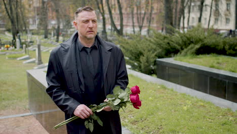 caucasian man in black raincoat and suit holding roses while walking in a graveyard