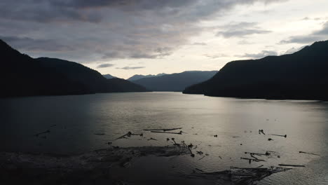 Fallen-tree-logs-poke-out-in-alpine-lake-at-entrance-of-river-with-soft-yellow-red-glow-in-sky-and-clouds