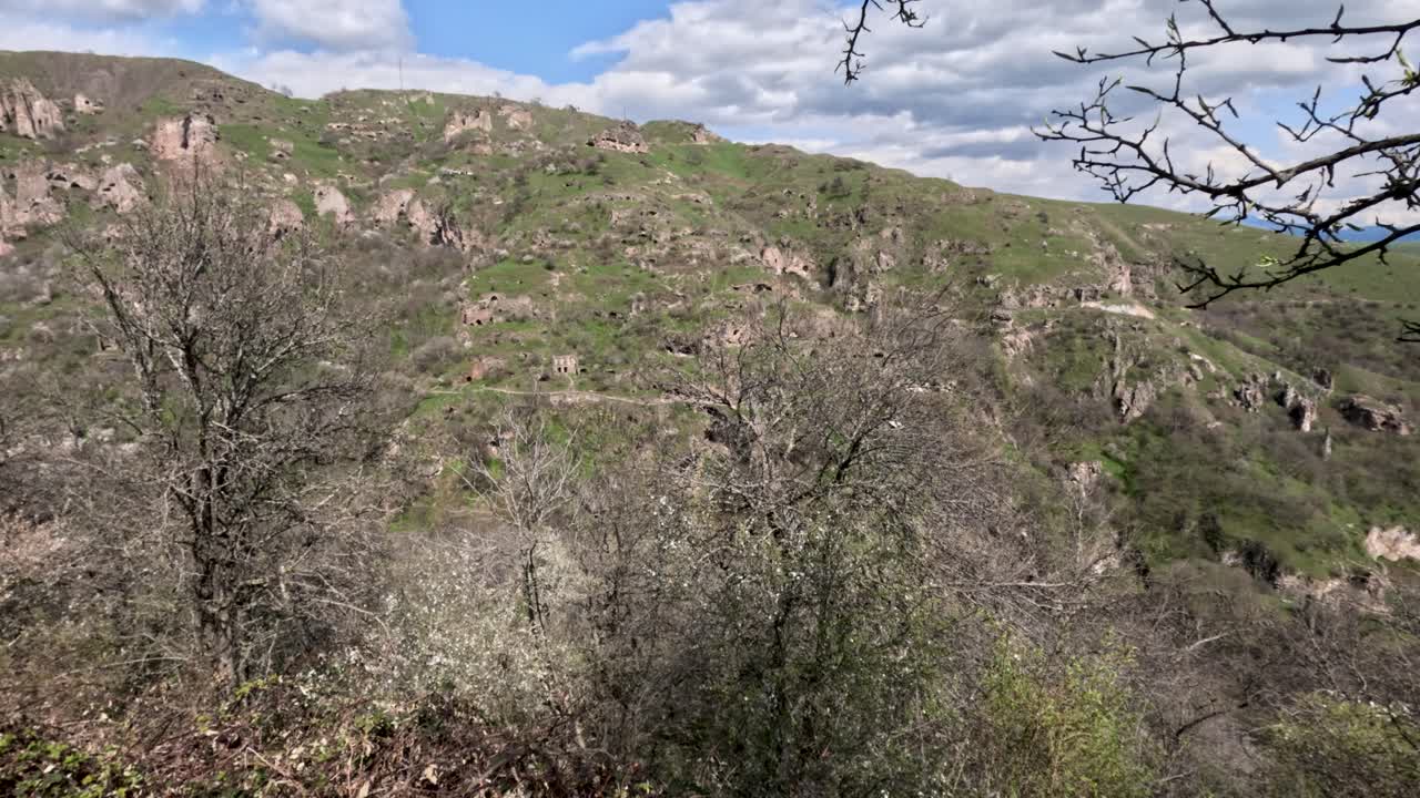 Uma vista mostra colinas ondulantes e vales com árvores esparsas. Nuvens brilhantes se movem pelo céu. Pássaros podem ser ouvidos ao fundo. O cenário é calmo e natural.