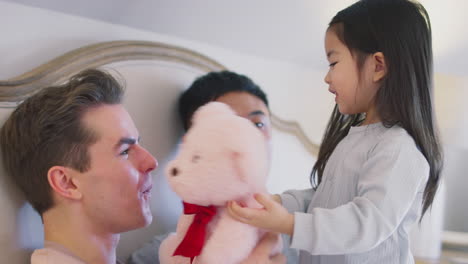 family with two dads in bed at home playing game with daughter and her soft toy