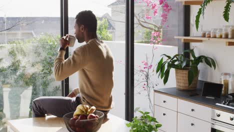 happy african american man sitting at countertop in kitchen, drinking coffee