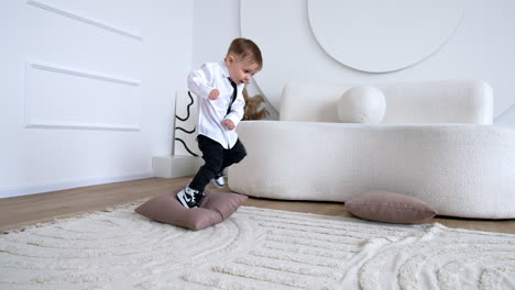 little kid in white shirt, black tie and jeans plays in the room. toddler plays and jumps by the cushions.